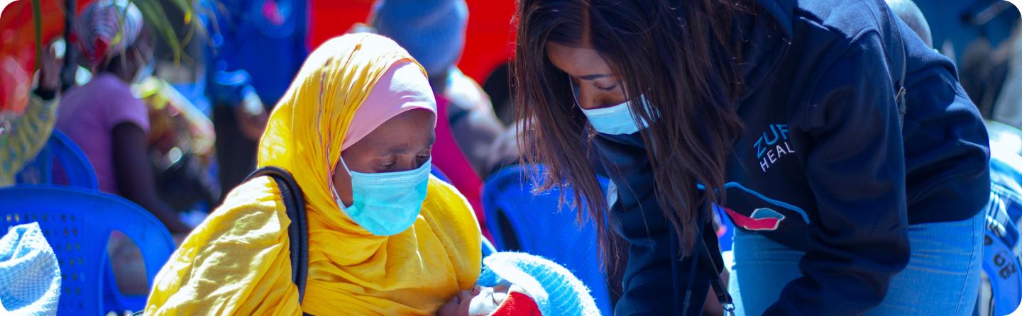 A masked Zuri Health nurse attending to a mother holding her baby at a community health camp